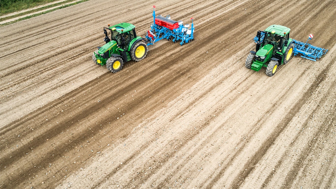 Δύο τρακτέρ John Deere 6M σε ένα γυμνό χωράφι. Το ένα έλκει τη φυτευτική μηχανή Monsomen.  Το άλλο έλκει συνδυασμό σπορείου Lemken