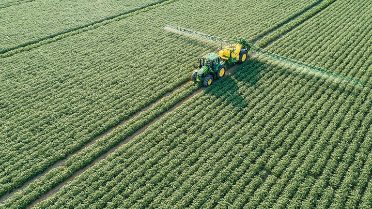 Τρακτέρ της σειράς John Deere 6M που έλκει ρυμουλκούμενο ψεκαστικό M700 σε χωράφι με καλλιέργεια πατάτας