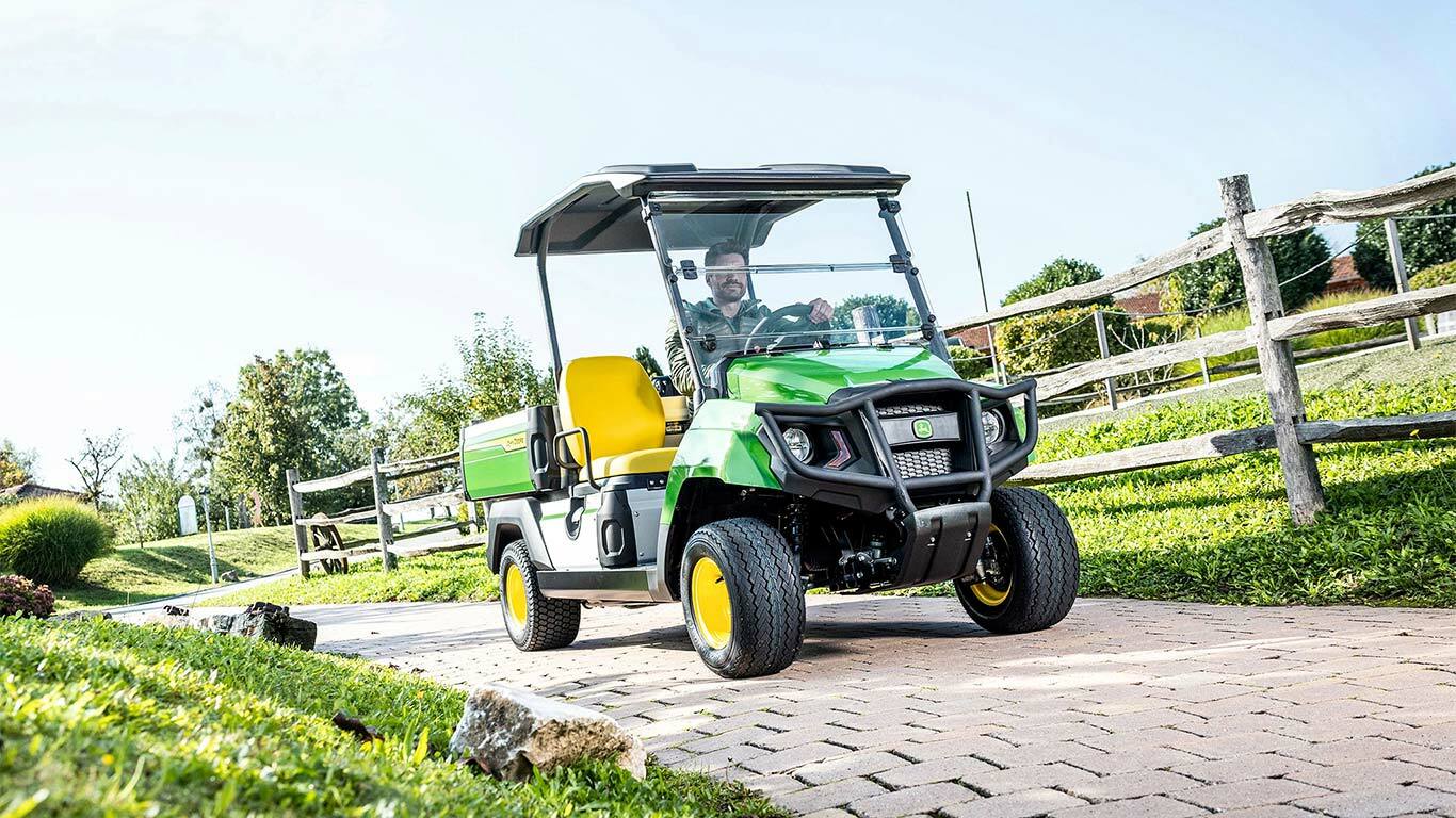 Πράσινο όχημα γενικής χρήσης Gator GS της John Deere που κινείται σε ασφαλτοστρωμένο δρόμο μέσα σε διαμορφωμένο χώρο με χόρτο και ξύλινους φράχτες.