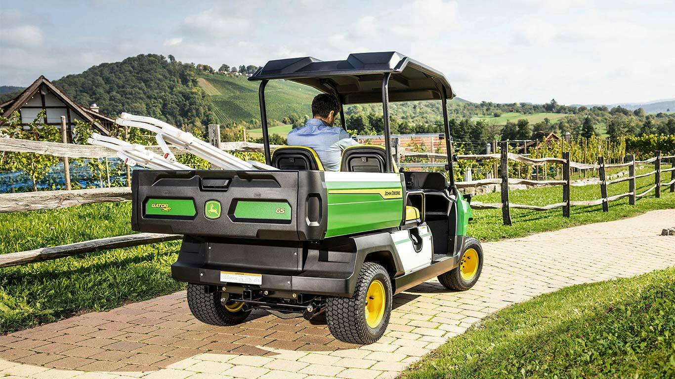 Πίσω όψη ενός Gator GS της John Deere που κινείται κατά μήκος μιας διαδρομής από τούβλα με γραφικούς λόφους και αμπελώνες στο φόντο.