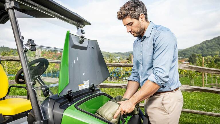 Άτομο που τοποθετεί έναν σάκο με επένδυση στον μπροστινό χώρο αποθήκευσης ενός πράσινου Gator GS της John Deere σταθμευμένου σε εξωτερικό χώρο κοντά σε ξύλινο φράχτη και αμπελώνα.