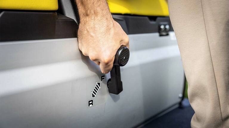 Κοντινό πλάνο ενός χεριού που χειρίζεται τον μοχλό αλλαγής ταχυτήτων σε Gator GS της John Deere, με ένδειξη της θέσης για μπροστινή ή προς τα πίσω κίνηση δίπλα στα κίτρινα καθίσματα.