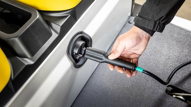 Χέρι που συνδέει ένα καλώδιο φόρτισης στην πλευρική θύρα ενός ηλεκτρικού οχήματος γενικής χρήσης Gator GS της John Deere.