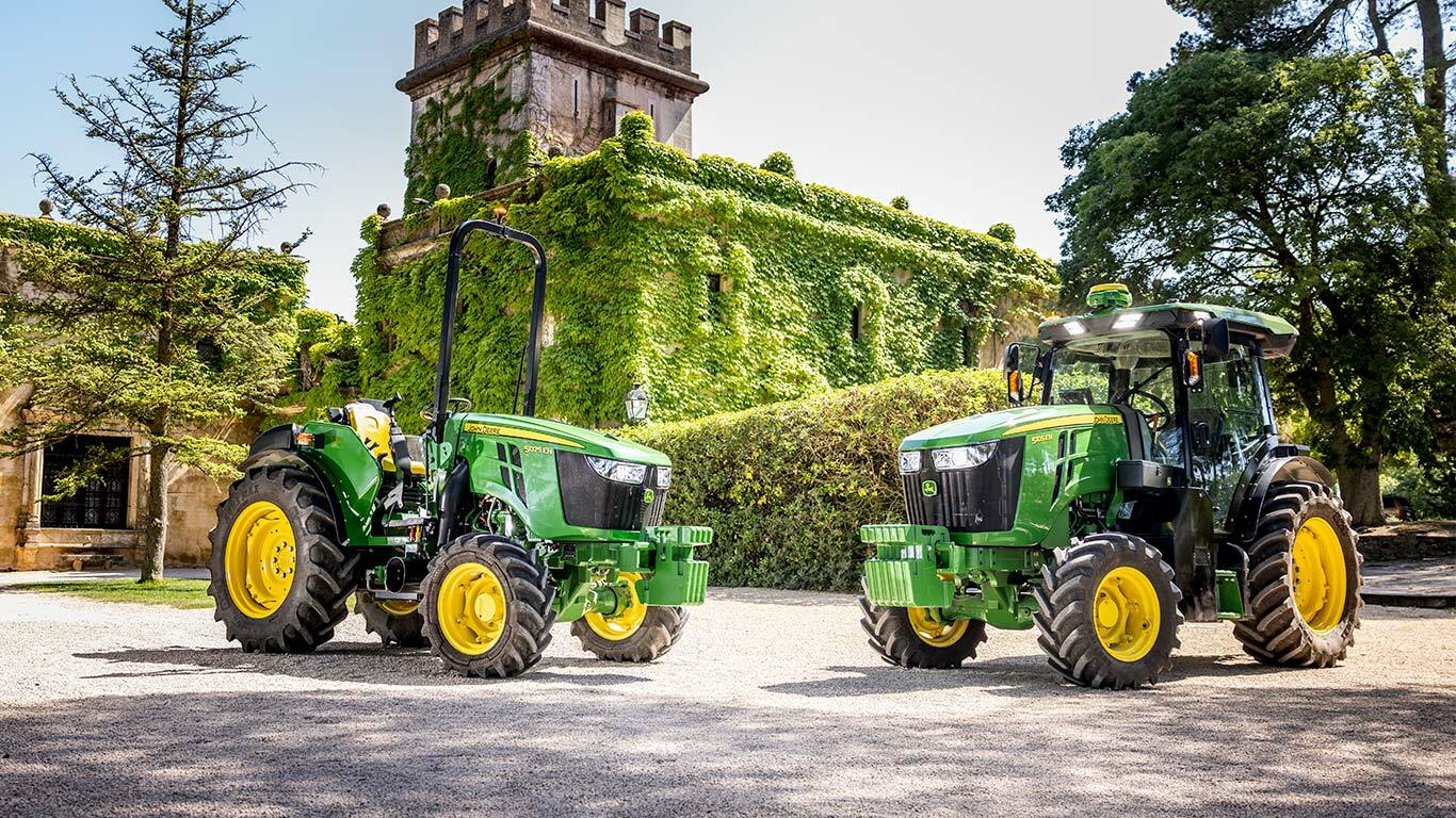 Πράσινο και κίτρινο τρακτέρ 5075EN της John Deere που διασχίζει έναν οπωρώνα σε χωματόδρομο, περιτριγυρισμένο από οπωροφόρα δέντρα και ηλιακό φως.