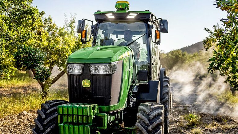 Πράσινο και κίτρινο τρακτέρ 5075EN της John Deere που διασχίζει έναν οπωρώνα σε χωματόδρομο, περιτριγυρισμένο από οπωροφόρα δέντρα και ηλιακό φως.