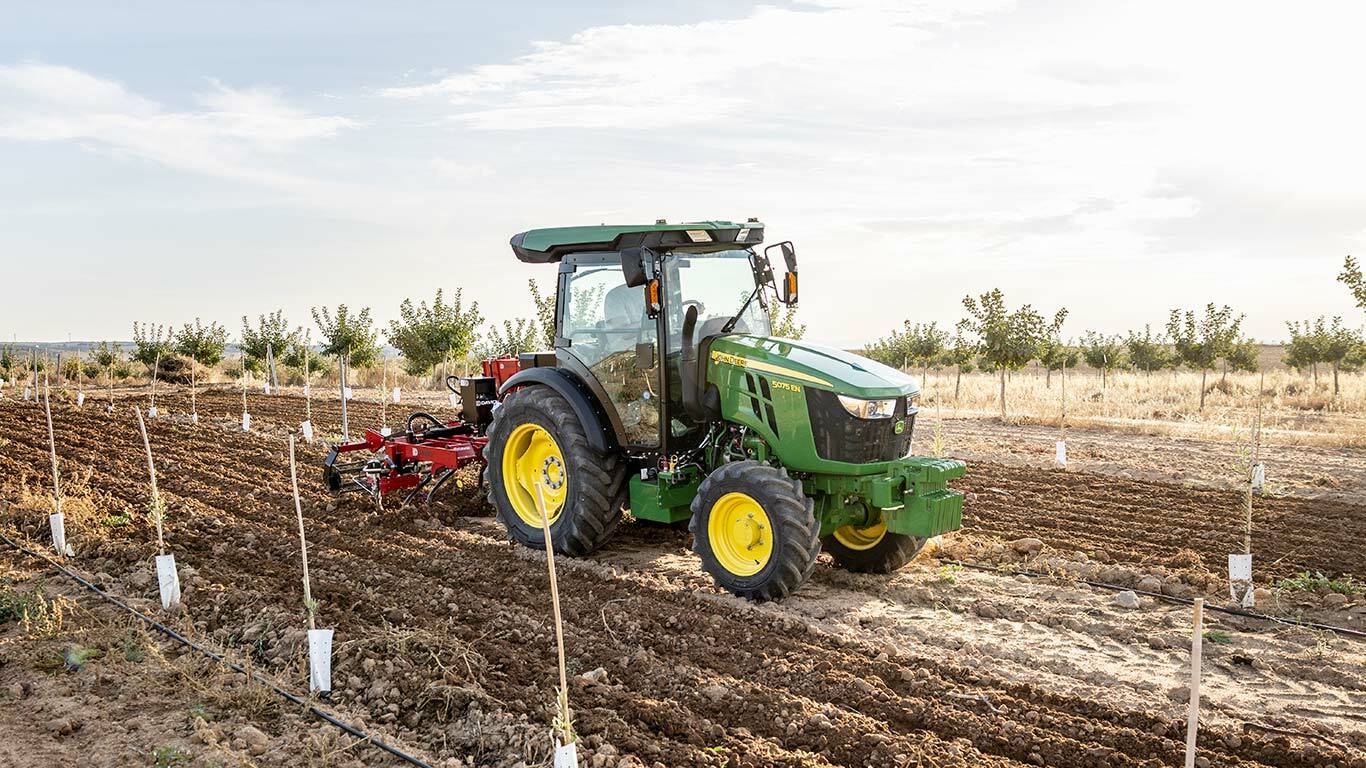Τρακτέρ John Deere 5075EN καλλιεργεί το έδαφος σε ανοιχτό χωράφι με καθαρό ουρανό και φυτεμένες σειρές.