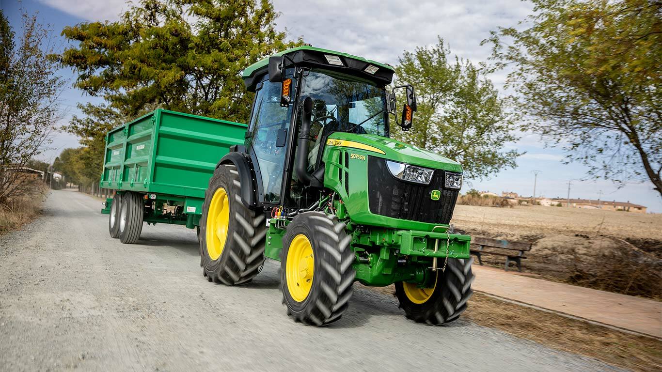 Τρακτέρ John Deere 5075EN τραβά πράσινο τρέιλερ σε αγροτικό χωματόδρομο με δέντρα στο φόντο.