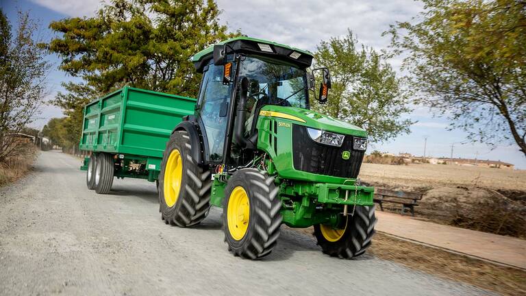 Τρακτέρ John Deere 5075EN τραβά πράσινο τρέιλερ σε αγροτικό χωματόδρομο με δέντρα στο φόντο.