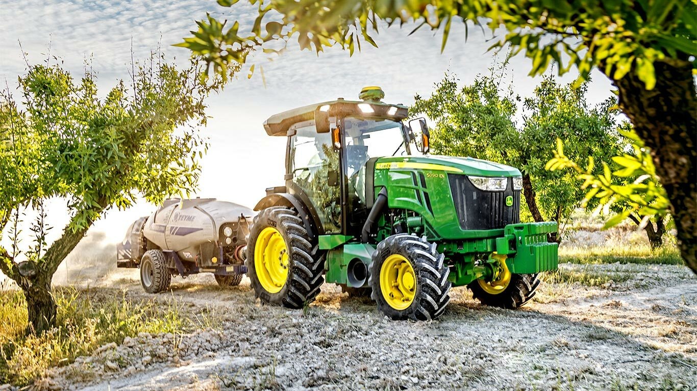 Πράσινο και κίτρινο τρακτέρ 5090EN της John Deere που ρυμουλκεί ένα ψεκαστικό σε έναν οπωρώνα μια ηλιόλουστη ημέρα.