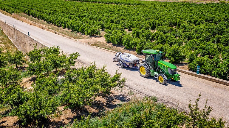 Πράσινο και κίτρινο τρακτέρ 5090EN της John Deere που ρυμουλκεί ένα ψεκαστικό σε ασφαλτοστρωμένο δρόμο ανάμεσα σε μεγάλες σειρές οπωρώνων κάτω από έναν καθαρό γαλάζιο ουρανό.