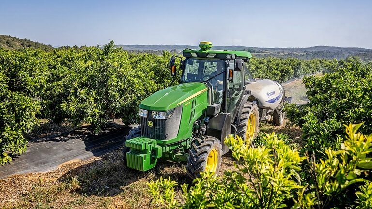 Πράσινο και κίτρινο τρακτέρ 5105EN της John Deere που ρυμουλκεί ένα ψεκαστικό σε έναν καταπράσινο οπωρώνα με λοφώδη εδάφη στο φόντο.