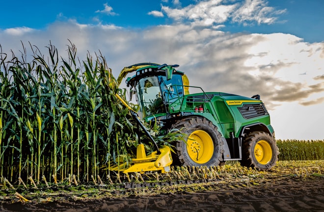Ενσιρωτικό μηχάνημα John Deere θερίζει καλαμπόκι σε χωράφι κάτω από γαλάζιο ουρανό με σύννεφα.