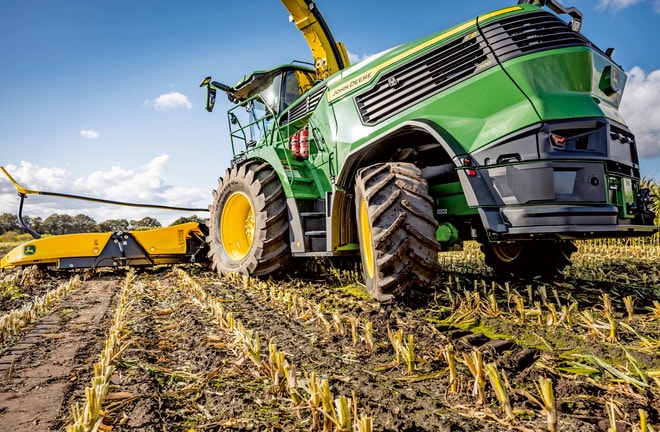 Ενσιρωτικό μηχάνημα John Deere που λειτουργεί σε θερισμένο χωράφι με καλαμπόκι, παρουσιάζοντας τα μεγάλα ελαστικά και τον μηχανισμό κοπής του μηχανήματος με φόντο έναν καταγάλανο ουρανό.